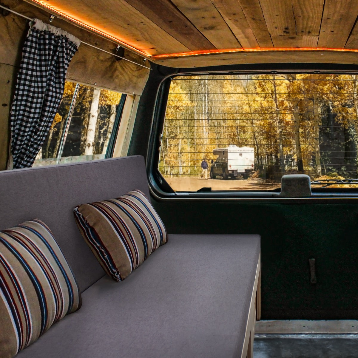 Cozy camper van interior featuring a gray bed with striped cushions, wooden ceiling, and large windows showcasing autumn scenery