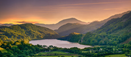 Lush green mountains surround a tranquil lake at sunset, with warm golden light illuminating the landscape and soft clouds above