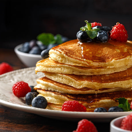 Fluffy golden pancakes stacked high, drizzled with syrup, topped with fresh blueberries, raspberries, and mint leaves on a rustic plate