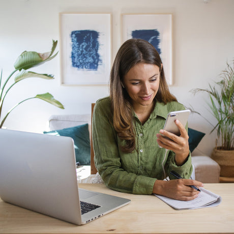 A woman in a green shirt smiles at her phone while sitting at a wooden desk with a laptop and notebook, surrounded by plants