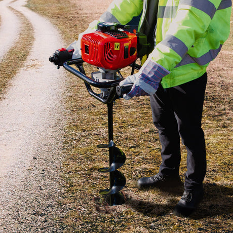 A person in a reflective jacket operates a red gas-powered auger on a gravel path, surrounded by dry grass and earth