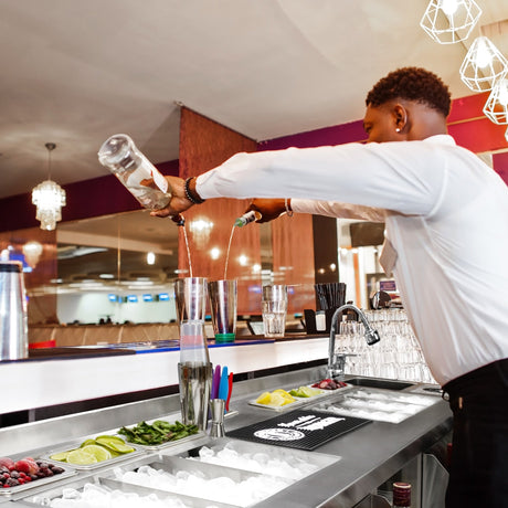 A bartender pours a colorful cocktail into a shaker, surrounded by fresh fruits, ice, and bar tools on a sleek metal counter