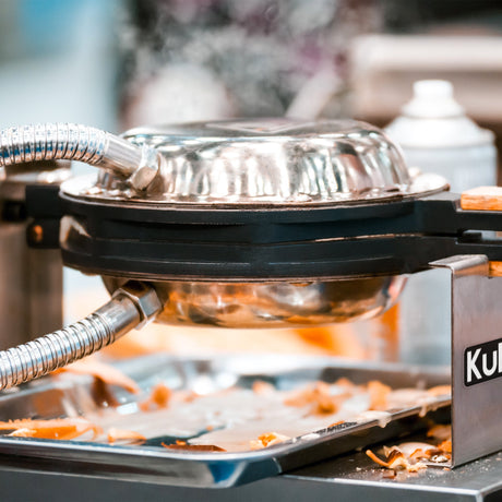 Shiny stainless steel waffle maker with steam rising, positioned on a countertop, surrounded by scattered waffle pieces and utensils