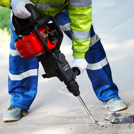 A worker in a bright green safety jacket and blue pants operates a red and black jackhammer on a concrete surface, creating dust and debris