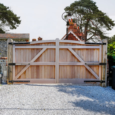 Sturdy wooden garden gate with a natural finish, featuring black metal hardware, set against a gravel driveway and lush greenery