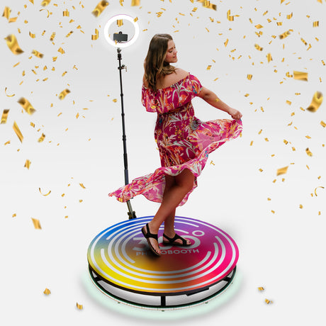 A woman in a vibrant pink floral dress twirls on a colorful photo booth platform, surrounded by golden confetti and soft lighting