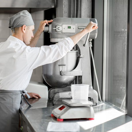 Chef in a white coat and gray apron adjusts a silver stand mixer on a stainless steel countertop, measuring ingredients in a clear cup
